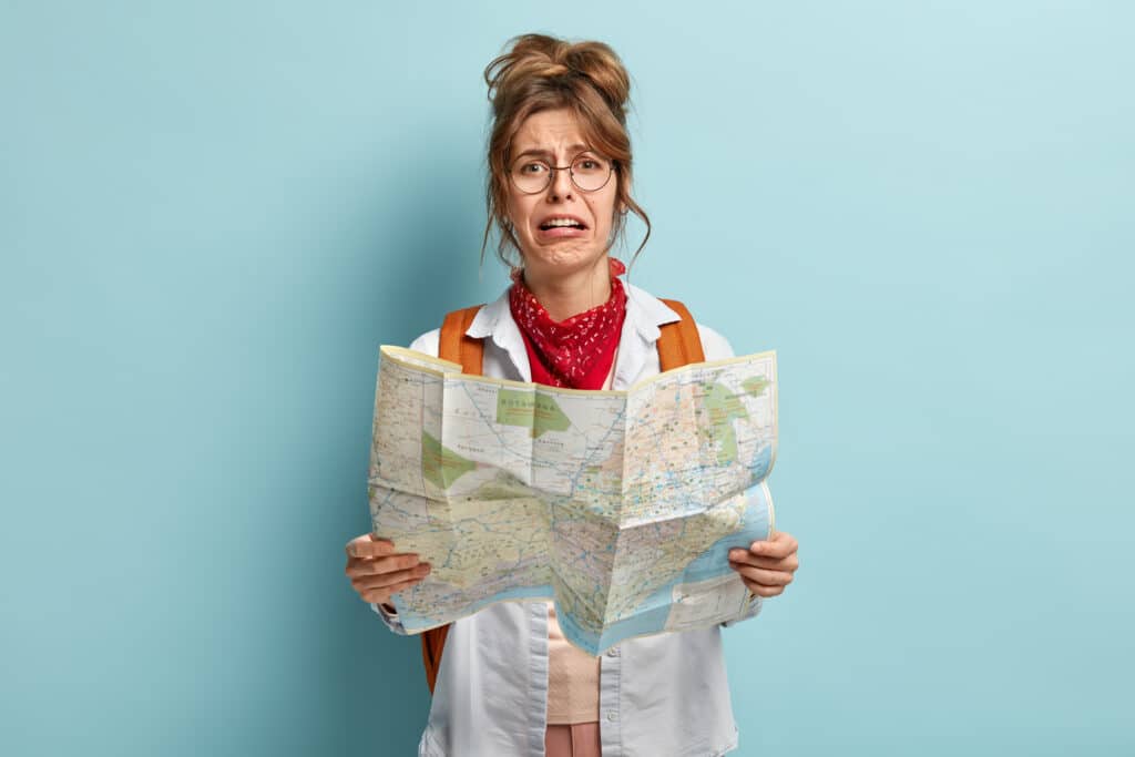 Crying female tourist lost in unknown destination, holds map, tries to find way, looks with dissatisfied dejected expression, wears round spectacles, has rucksack with packed things, stands indoor