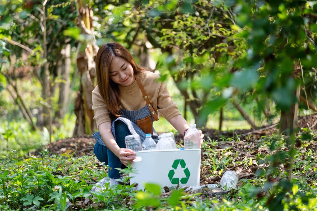Eco-friendly Travel Tips. A beautiful young asian woman collecting and putting plastic bottles into a recycle bin in the outdoors.