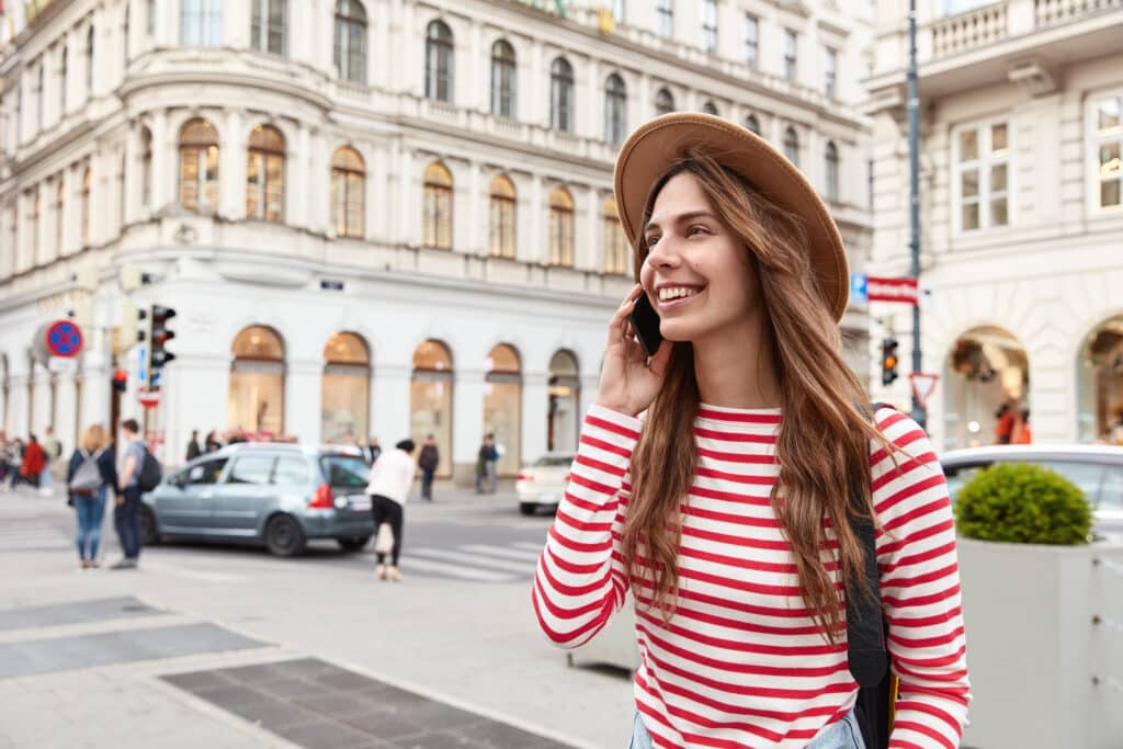 Essential Solo Travel Resources. Cheerful female wanderer strolls in city, enjoys communication, keeps modern smartphone near ear, focused aside, wears hat and striped jumper, explores streets, uses good internet connection