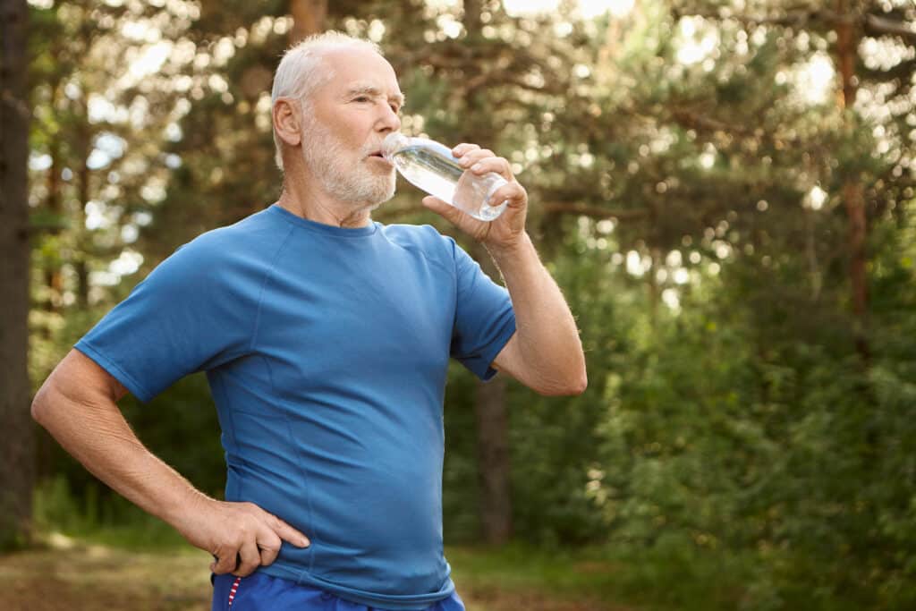 Portrait of attractive active male pensioner with bald head and stubble refreshing himself after jogging outdoors, standing against pine forest background, holding bottle of drinking water
