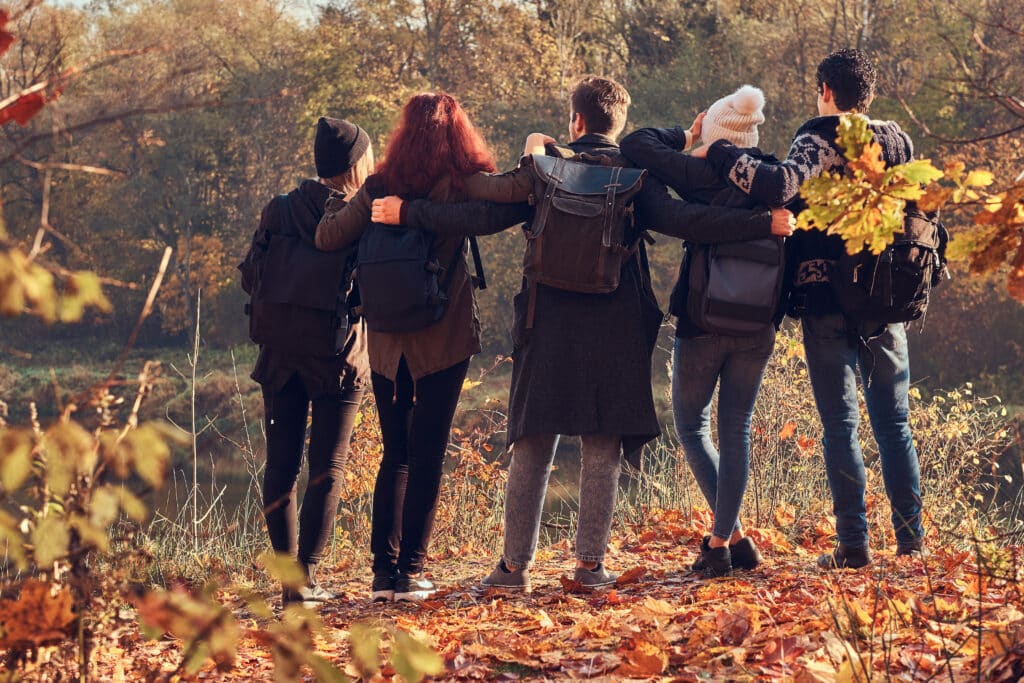 Travel, hiking, adventure concept. Back view of young friends hugging together and looking at the lake in beautiful autumn forest.