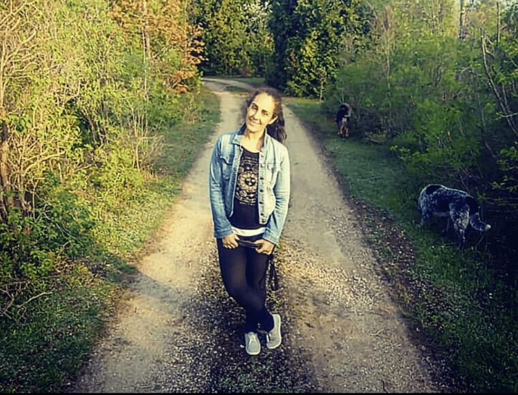 A woman with long brown hair wearing a jean jacket standing in the middle of a gravel trail in a forest.