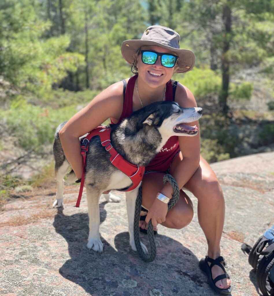 A woman in a hat, sunglasses, and sandals crouches down on a large rock to hug a large dog. They are outside in the woods.